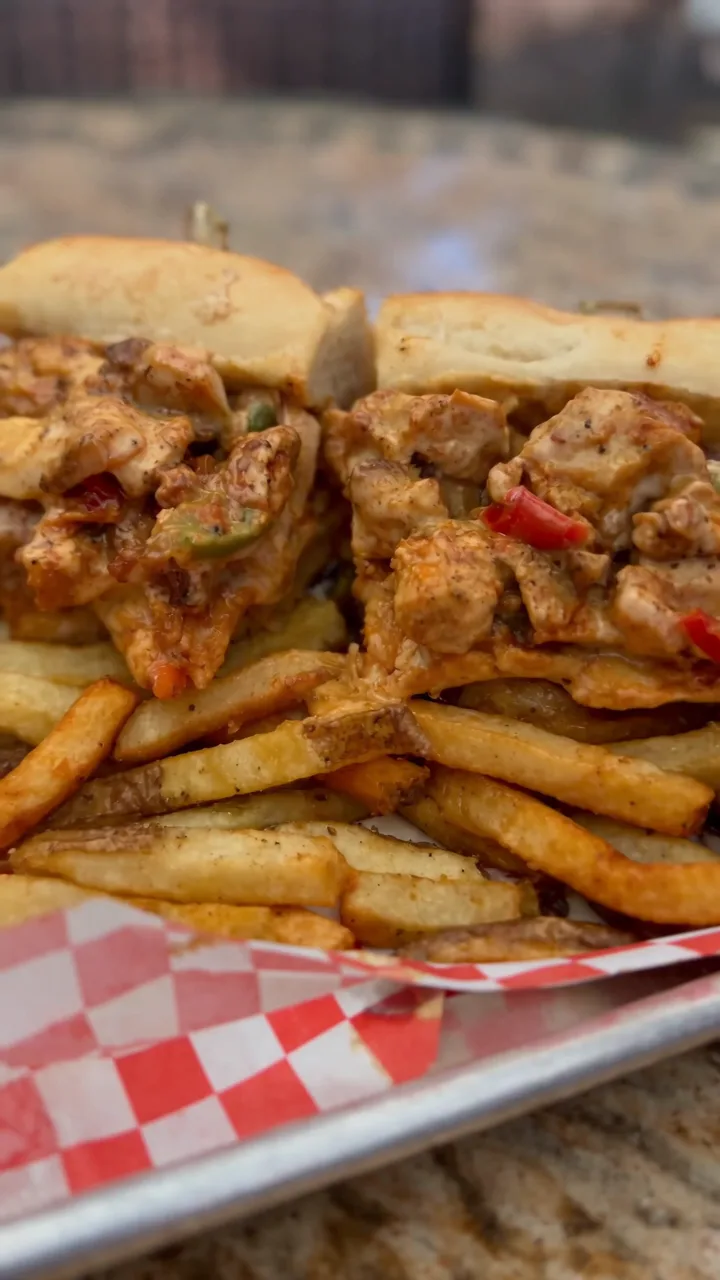 Generated view of a past Chicken Cheesesteak special: chicken, peppers and jalapenos on a hoagie roll over fries in a red-checkered tray, with white sauce poured over it.