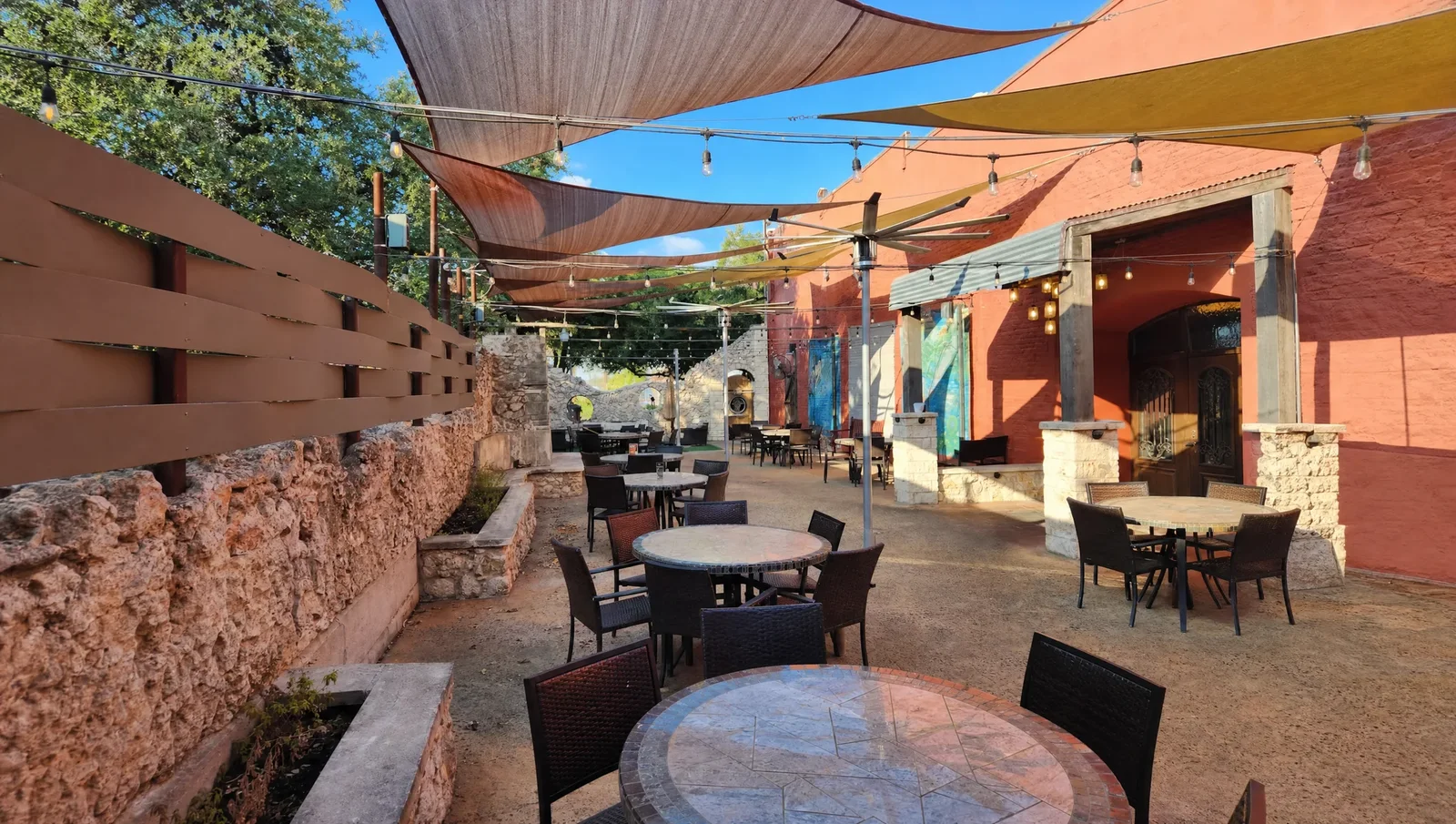 A wide interpretation of The Victory Tap’s real patio in afternoon light, with mosaic tables and shade sails.